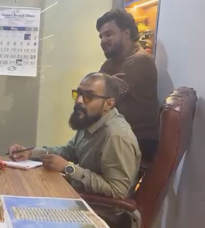Sumit Choudhari seated writing at a desk with Vinodkumar Prajapati (Vicky) standing behind him